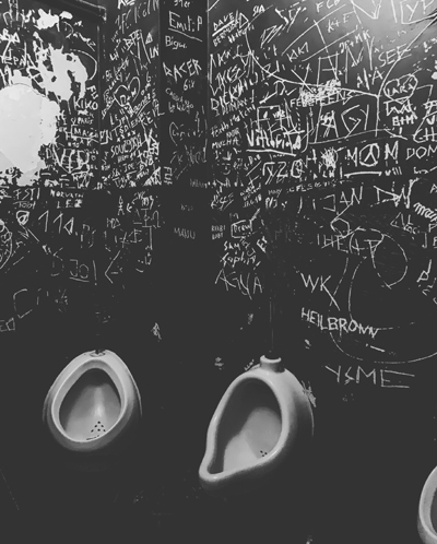Schwarze Wand mit weißen Graffitischriftzügen und zwei weißen Urinalen. 