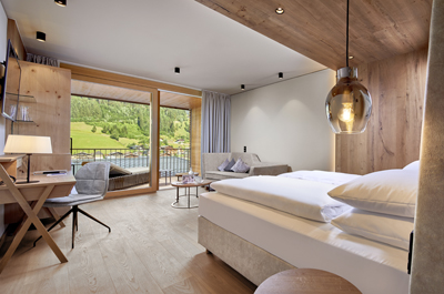 Vom Bett aus fällt der Blick – neben Schreibtisch, Sofa und Schrank – auf den Balkon und das ländliche Panorama. © DAS EDELWEISS Salzburg Mountain Resort, Fotograf Michael Huber